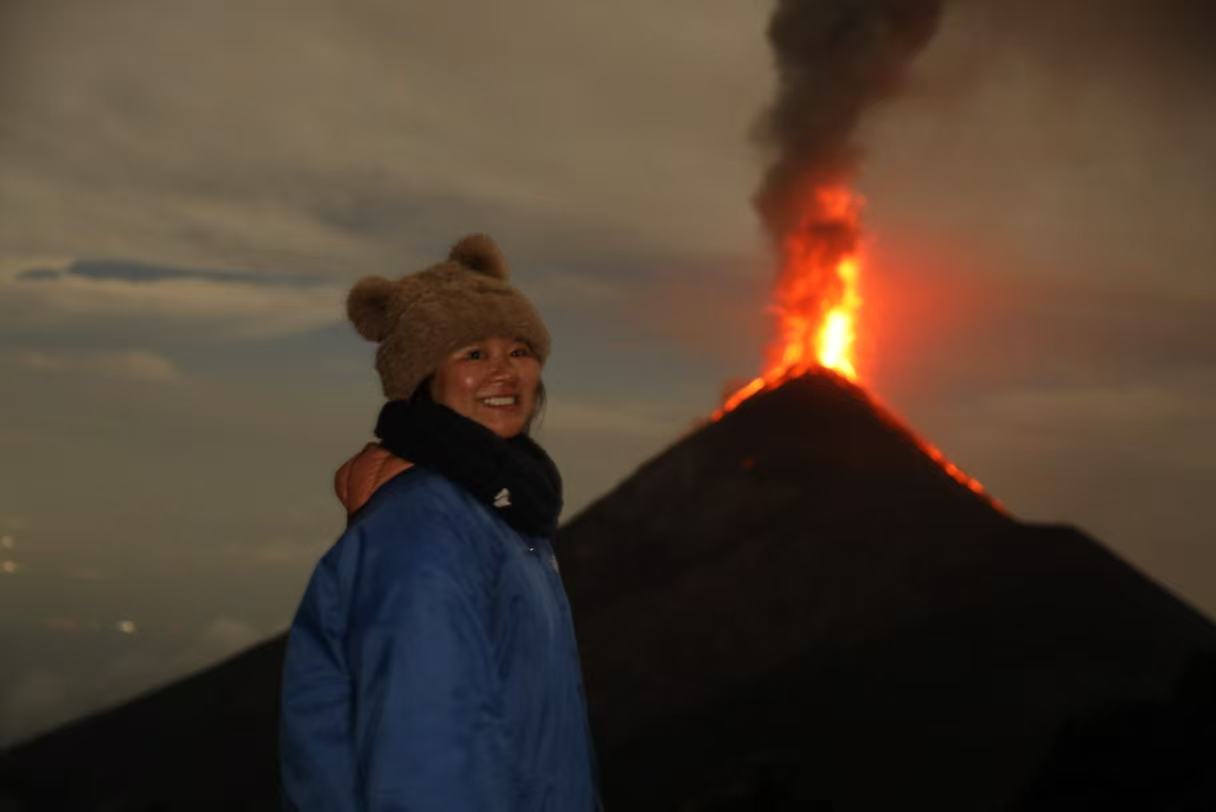 Jeanie smiling on a trip, with an erupting volcano behind her.